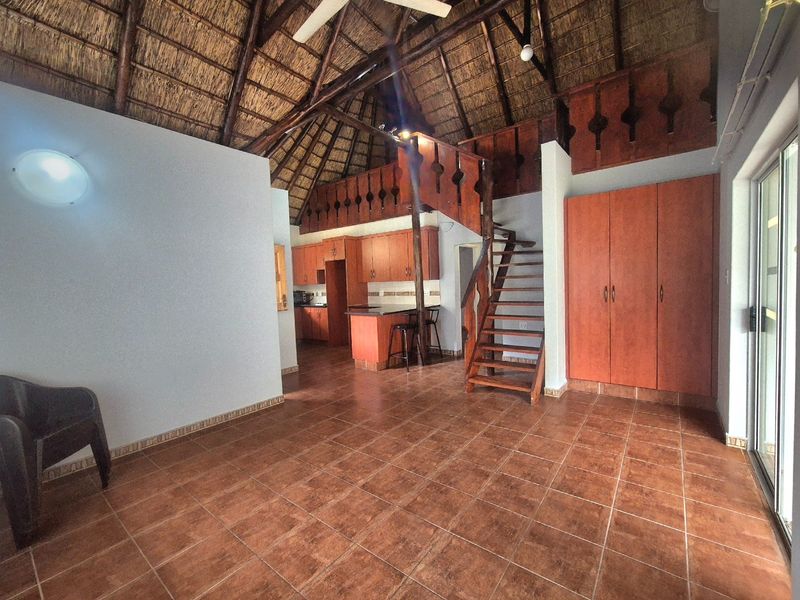 Open-plan living area featuring a high thatched roof with exposed timber trusses, tiled flooring, built-in wooden cabinetry, and a wooden staircase to a loft.