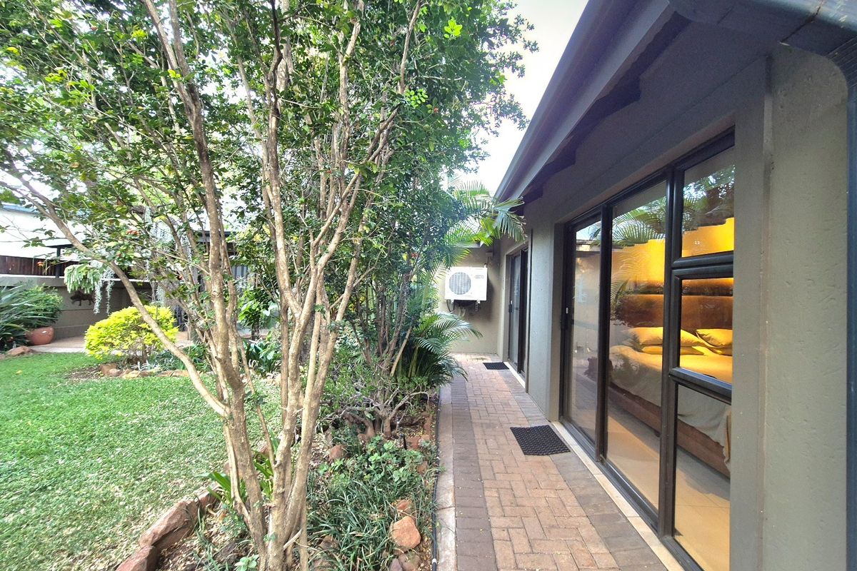 A well-maintained rear exterior view featuring a paved walkway alongside the house, mature garden landscaping, and sliding glass doors providing access to the interior.