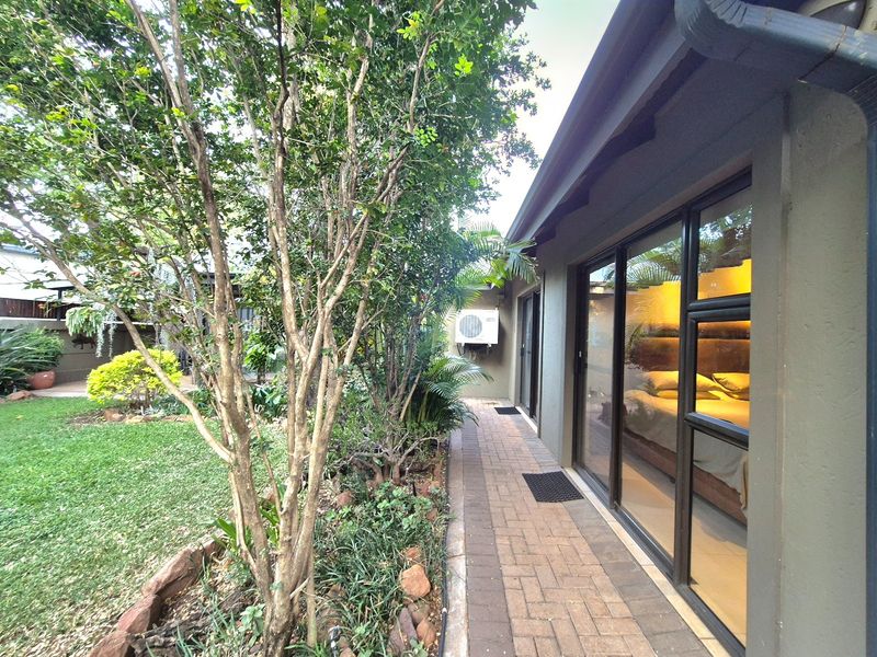A well-maintained rear exterior view featuring a paved walkway alongside the house, mature garden landscaping, and sliding glass doors providing access to the interior.
