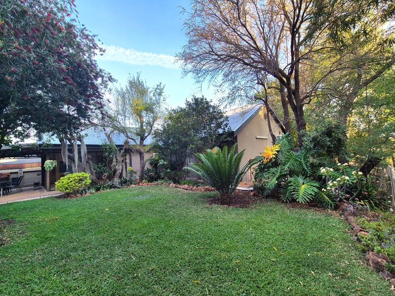 A lush, well-maintained garden featuring a manicured lawn, mature trees, and a paved patio area adjacent to the main house structure.