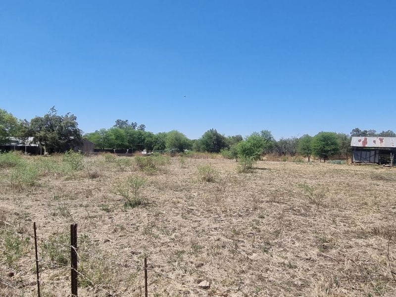 A wide-angle view of a vacant residential plot featuring dry grass, perimeter wire fencing, mature trees, and a small, weathered outbuilding.