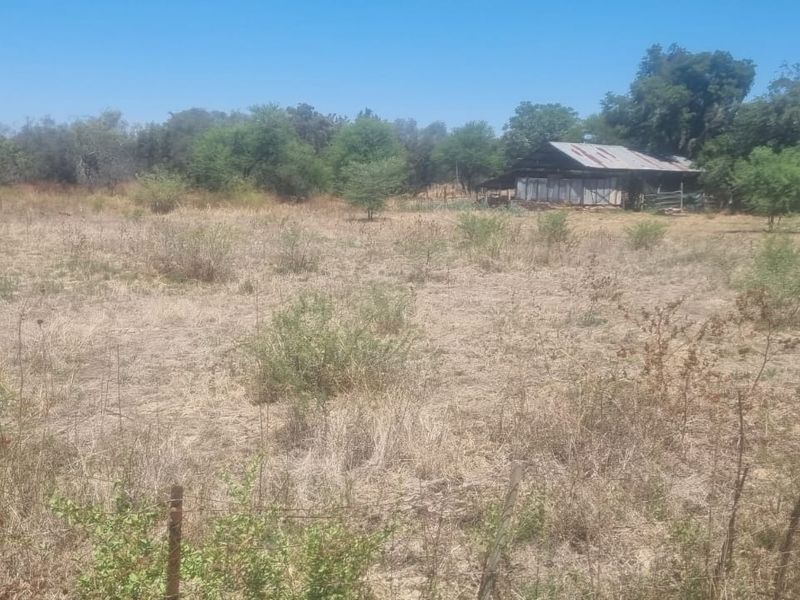 A view of a vacant residential plot featuring dry grass, mature trees, wire fencing, and an existing outbuilding structure on the property.