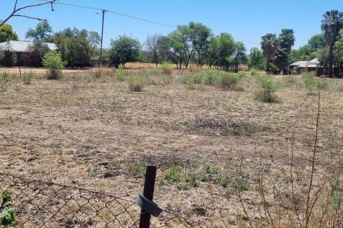 A vacant residential plot featuring natural vegetation, wire perimeter fencing, and surrounding mature trees in a quiet neighborhood setting.