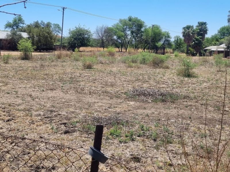 A vacant residential plot featuring natural vegetation, wire perimeter fencing, and surrounding mature trees in a quiet neighborhood setting.
