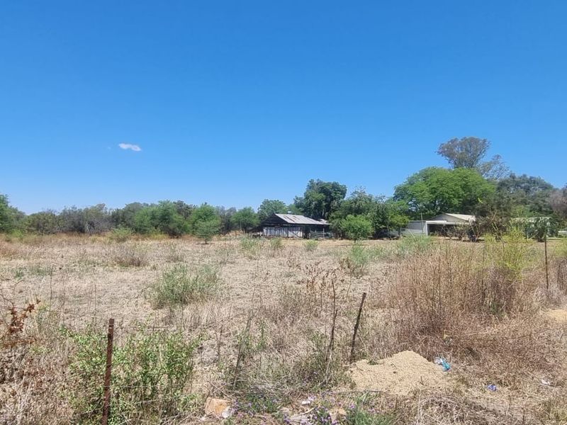 A view of a vacant residential plot featuring dry vegetation and perimeter wire fencing under a clear blue sky.