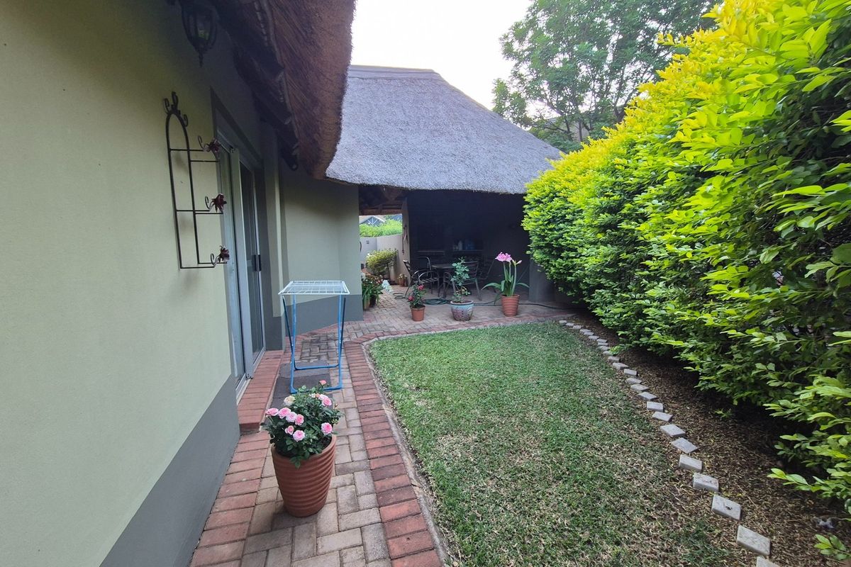 A well-maintained rear garden featuring a brick-paved patio, lush green lawn, and a covered outdoor area with a traditional thatched roof.