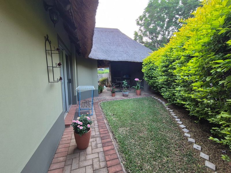 A well-maintained rear garden featuring a brick-paved patio, lush green lawn, and a covered outdoor area with a traditional thatched roof.