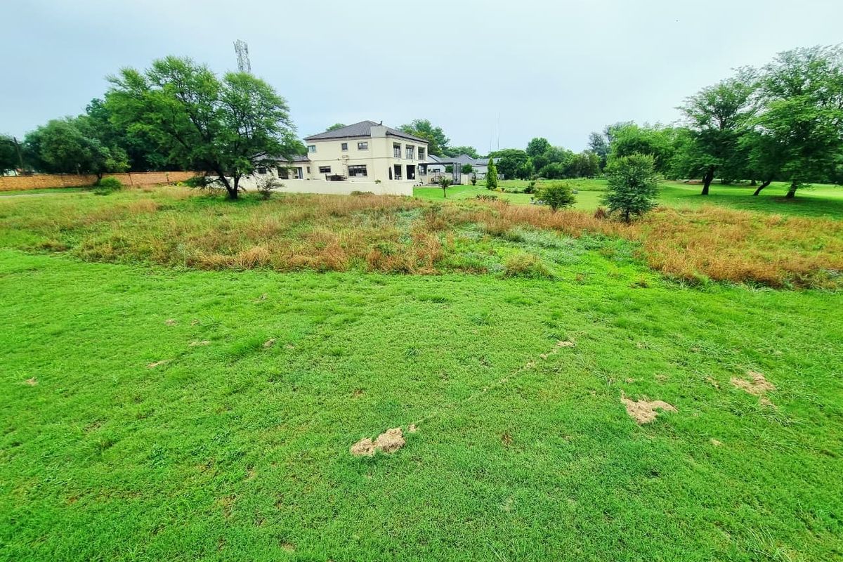A wide-angle view of a residential vacant land plot featuring a grassy lawn, mature trees, and a boundary wall with neighboring houses in the background.
