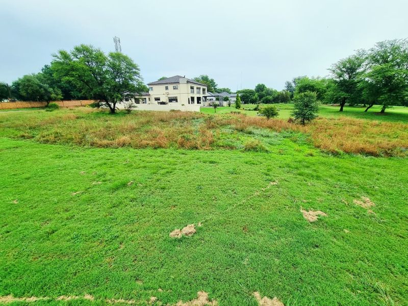 A wide-angle view of a residential vacant land plot featuring a grassy lawn, mature trees, and a boundary wall with neighboring houses in the background.