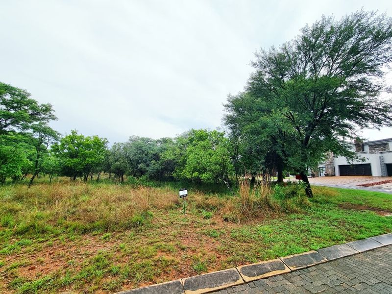 A vacant residential plot featuring established trees and natural vegetation, adjacent to a paved driveway and existing residential infrastructure.