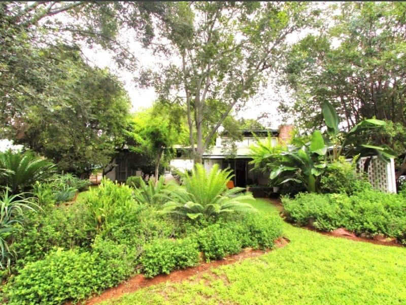 A lush, mature garden featuring established trees, vibrant greenery, and a well-maintained lawn leading toward the rear elevation of the house.