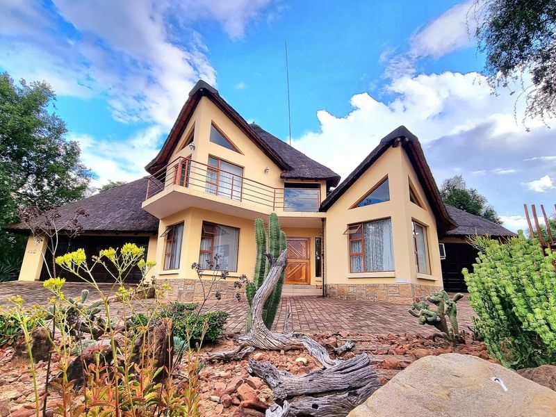 A well-maintained double-story house featuring a traditional thatch roof, stone accents, a private balcony, and a paved driveway with double garage access.
