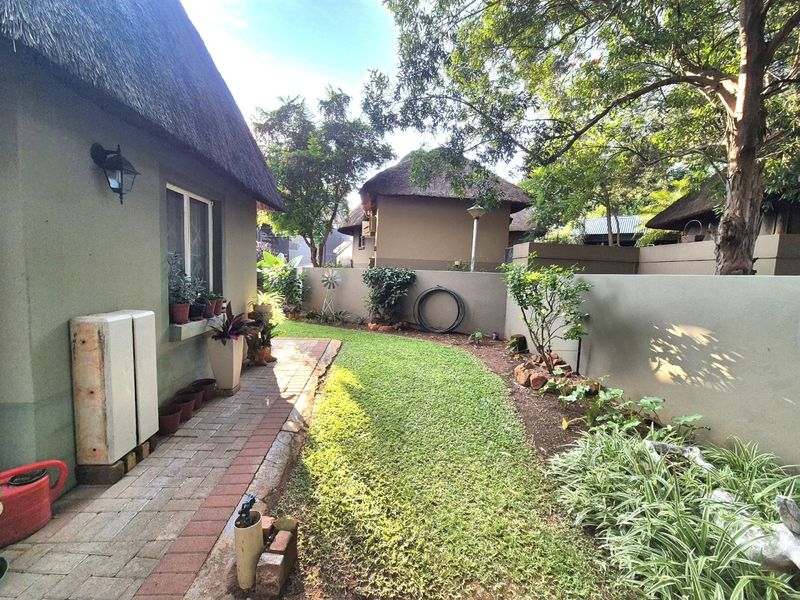 A well-maintained rear garden area featuring a brick paved pathway, lush lawn, and boundary wall, adjacent to a house with a traditional thatch roof.