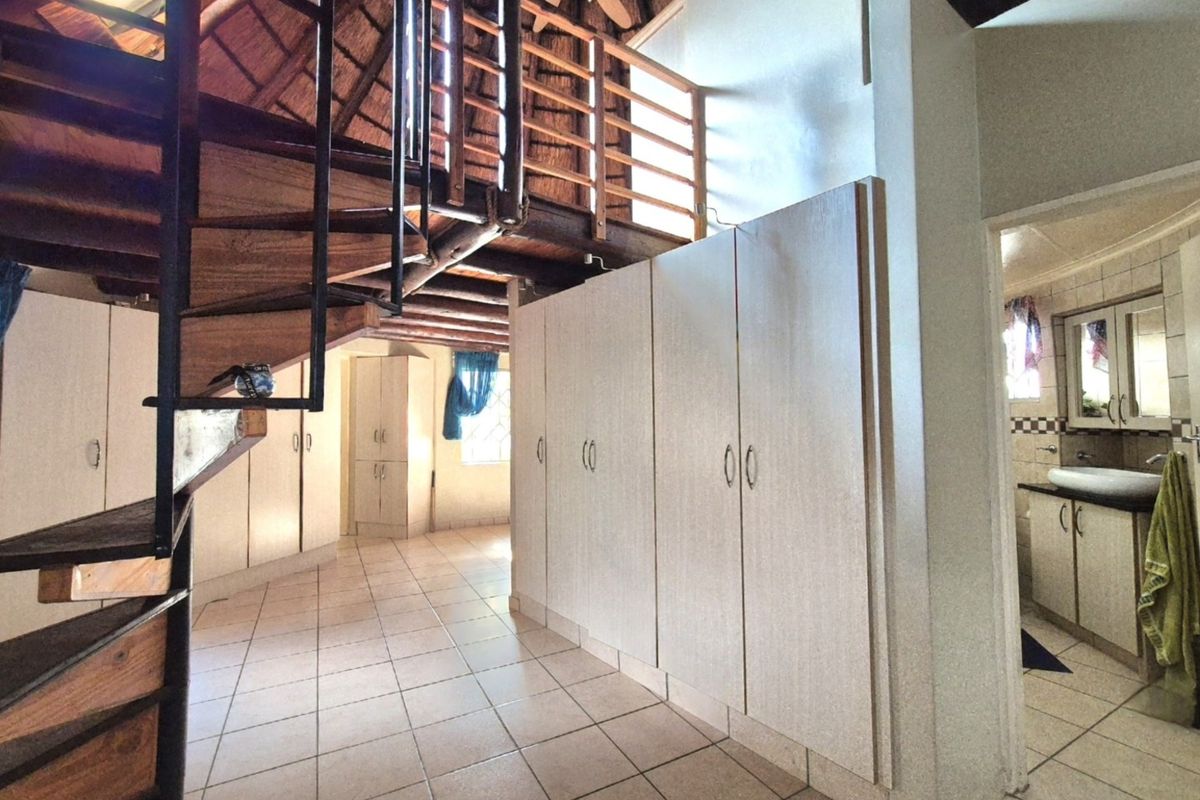 Spacious bedroom featuring a spiral staircase, extensive built-in cupboards, tiled flooring, and an open-plan en-suite bathroom under a high thatch ceiling.