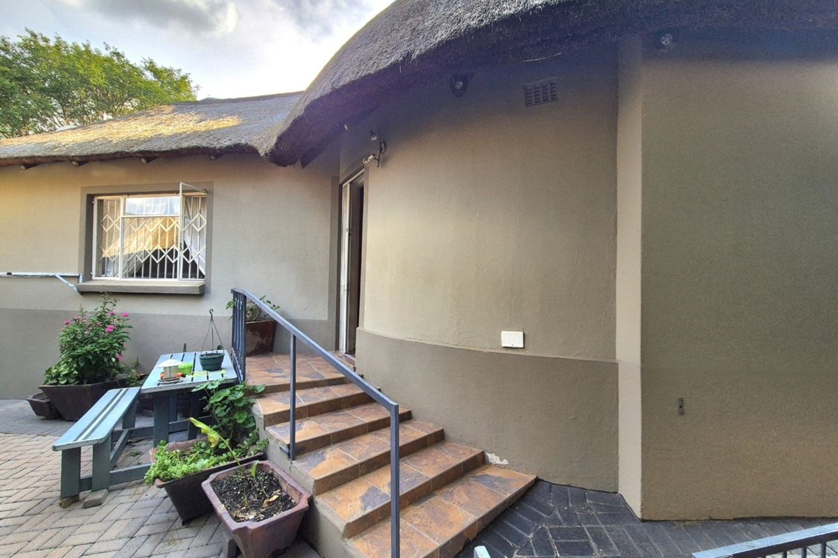 Rear exterior view of a house featuring a traditional thatch roof, tiled entrance steps with a metal handrail, and paved patio area.