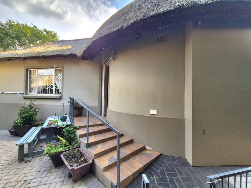 Rear exterior view of a house featuring a traditional thatch roof, tiled entrance steps with a metal handrail, and paved patio area.