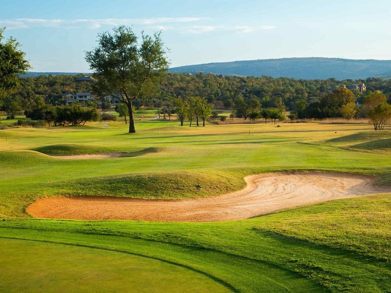 A scenic view of a well-maintained golf course featuring rolling green fairways and a sand bunker with distant hills in the background.