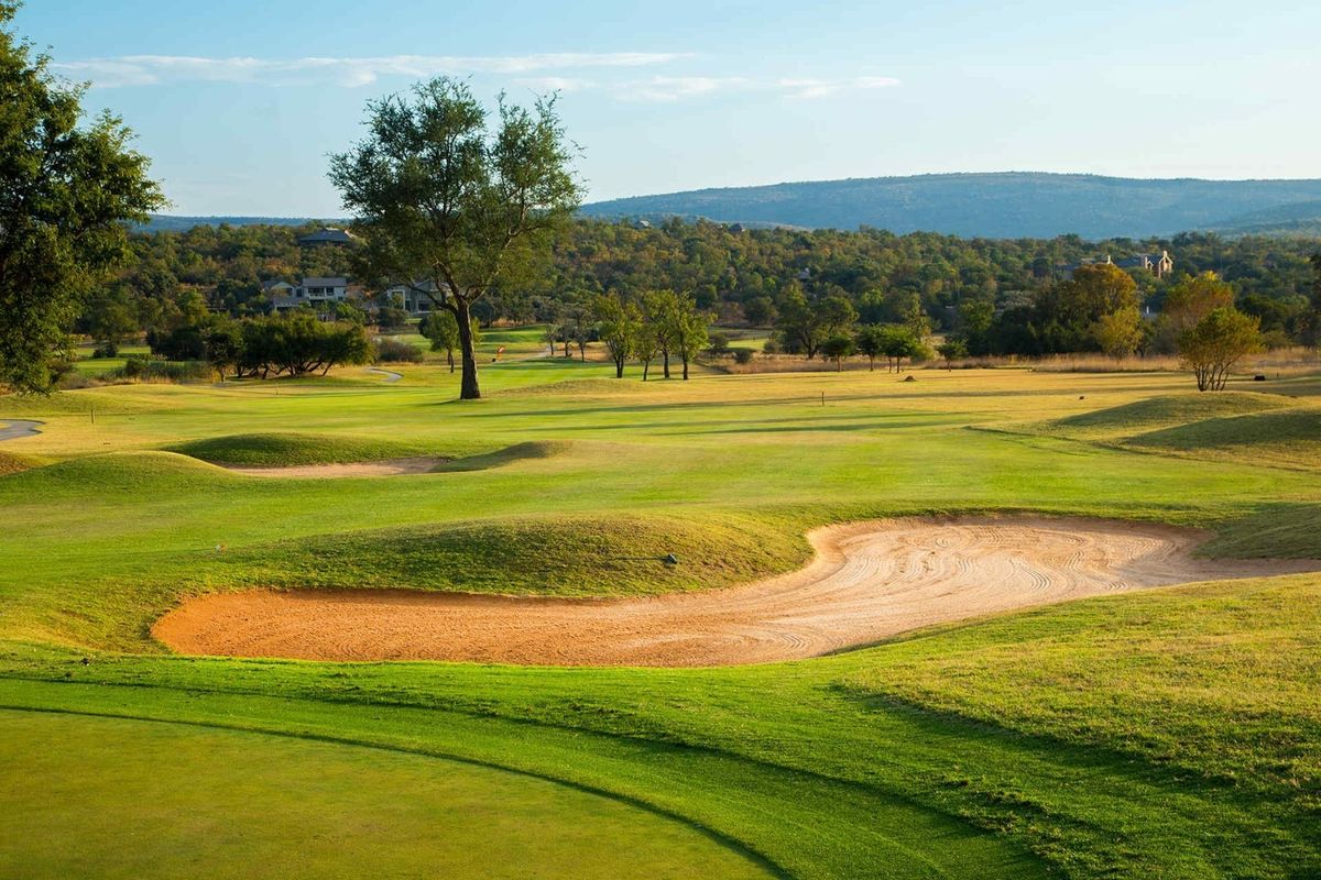 A scenic view of a well-maintained golf course featuring rolling green fairways and a sand bunker with distant hills in the background.