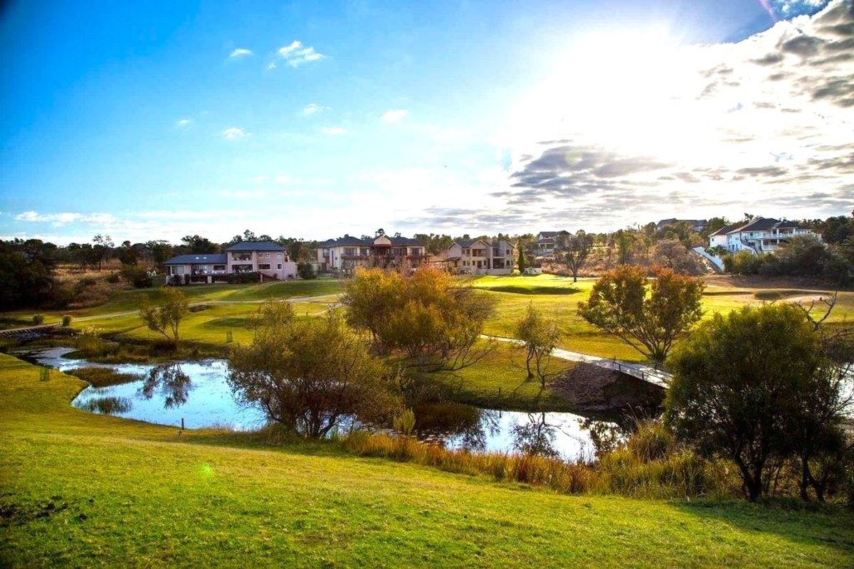 A scenic view of the residential estate grounds featuring manicured lawns, a natural water feature, and a pedestrian bridge.