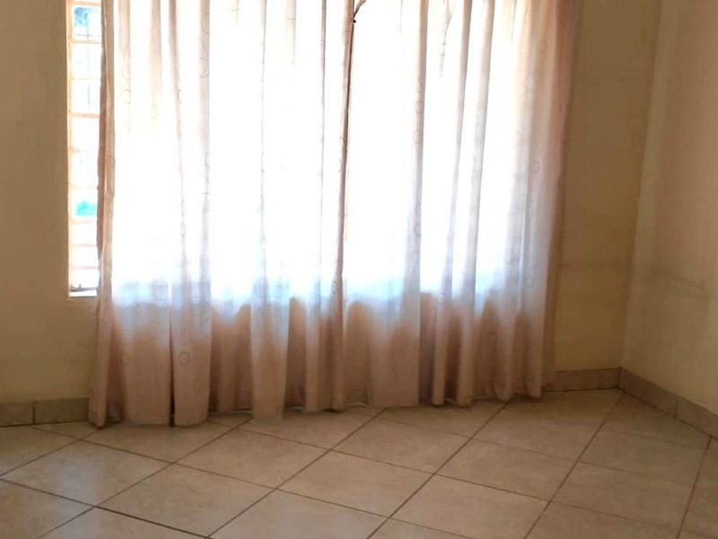 An empty bedroom featuring tiled flooring and a window with curtains, showing signs of wear on the walls.
