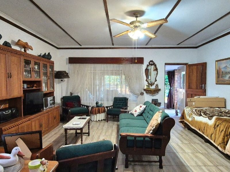 A spacious living room featuring tiled flooring, timber ceiling beams, and a central ceiling fan, offering a traditional layout with direct access to the exterior.