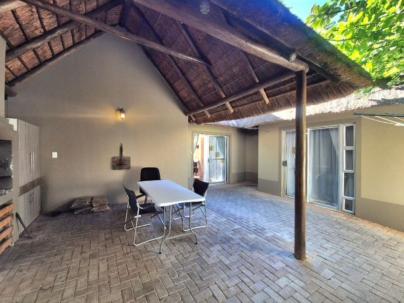 A covered outdoor patio featuring a traditional thatched roof, timber support posts, brick paving, and a built-in braai area with direct access to the home.