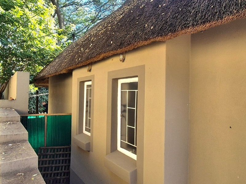 A side view of a house featuring a traditional thatched roof, plastered exterior walls, and white-framed casement windows set within a garden area.