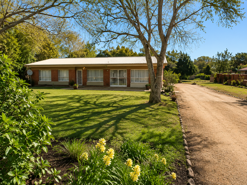 A single-story brick house with a corrugated metal roof, featuring a large front lawn, mature trees, and a gravel driveway.