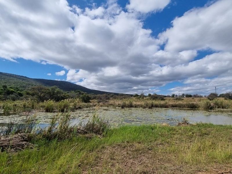 A scenic view of a natural pond or wetland area surrounded by grassy banks and rolling hills under a cloudy sky.