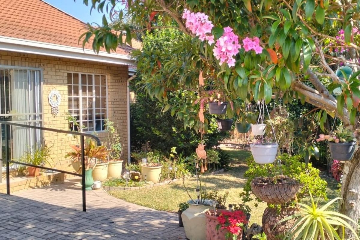 A view of the rear brick facade featuring a paved patio area, metal handrail, and a well-established garden with mature trees.