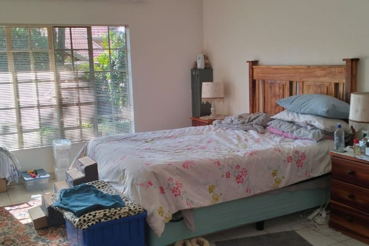 A functional bedroom featuring tiled flooring and large windows with venetian blinds, offering natural light and a view of the garden.