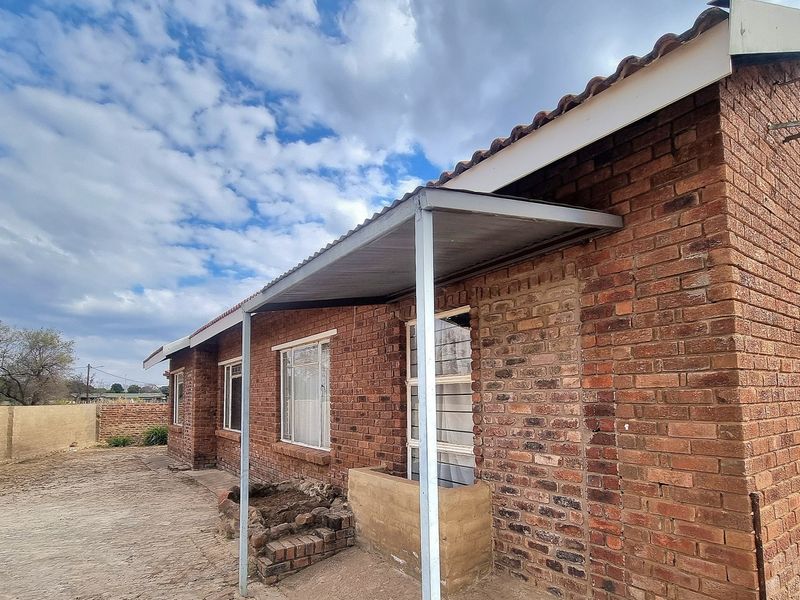 Rear exterior view of a brick house featuring a simple corrugated metal patio cover, concrete paving, and a small built-in brick planter box.