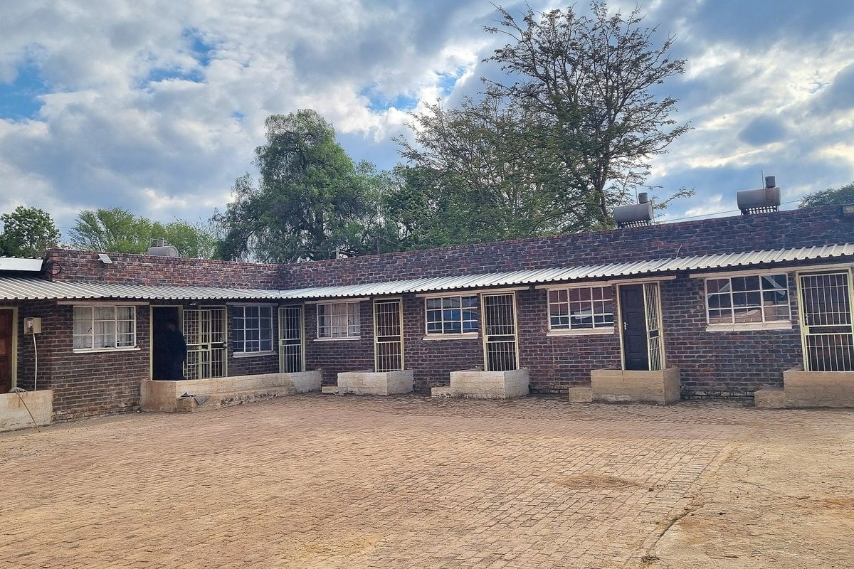 Rear view of a face brick residential building featuring multiple entrances with security gates, corrugated roofing, and a paved courtyard area.