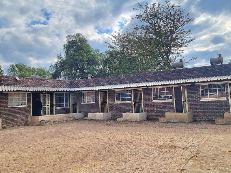 Rear view of a face brick residential building featuring multiple entrances with security gates, corrugated roofing, and a paved courtyard area.