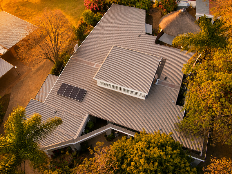 Aerial view of the property showcasing the corrugated metal roof, integrated solar panels, surrounding garden, and a detached thatch roof gazebo.
