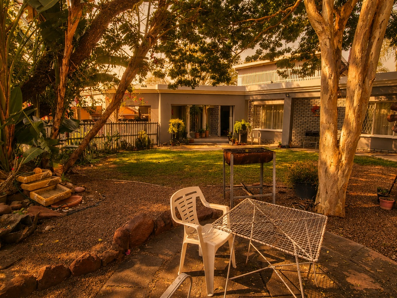 A spacious rear garden featuring a stone-paved patio area, mature trees, and a view of the house's rear facade with security features.