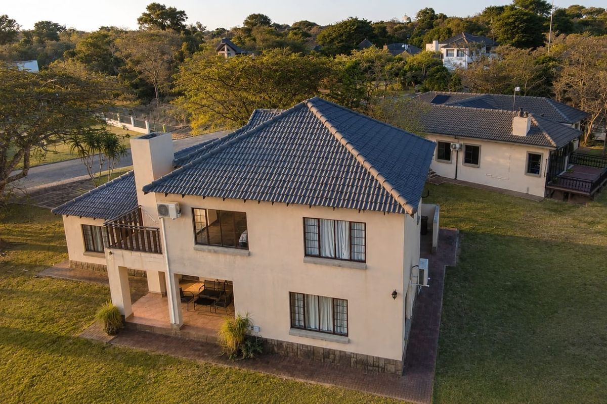 Aerial view of a two-story residential house featuring a tiled roof, chimney, balcony, and a paved patio area set within a landscaped garden.