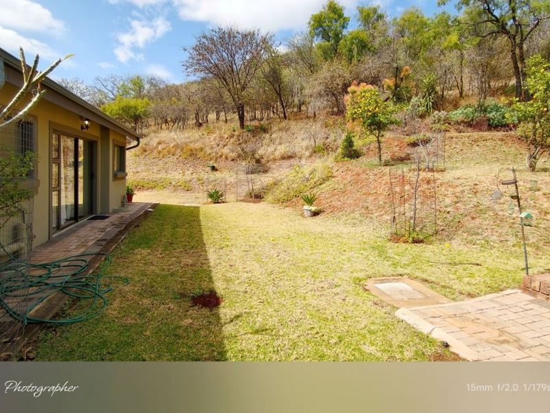 A rear exterior view of the property featuring a paved patio area, sliding glass doors, and a sloped garden with established trees.
