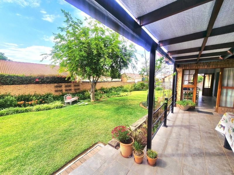 A well-maintained covered patio overlooking a lush green garden with a brick boundary wall and established trees.