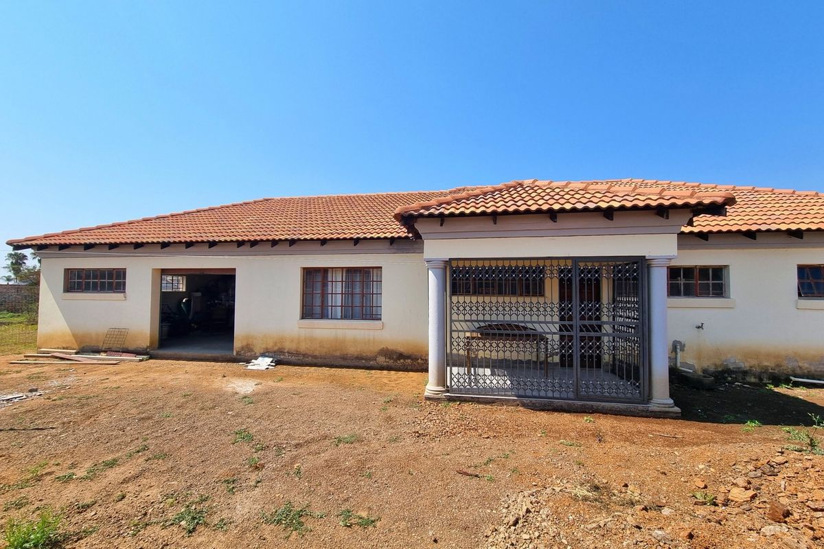 Rear view of a single-story house featuring a covered patio with security gate, terracotta roof tiles, and an open garage area with unfinished surrounding grounds.