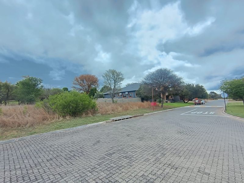 A street-level view of the residential property showing the paved road, stormwater drainage, and surrounding landscape.