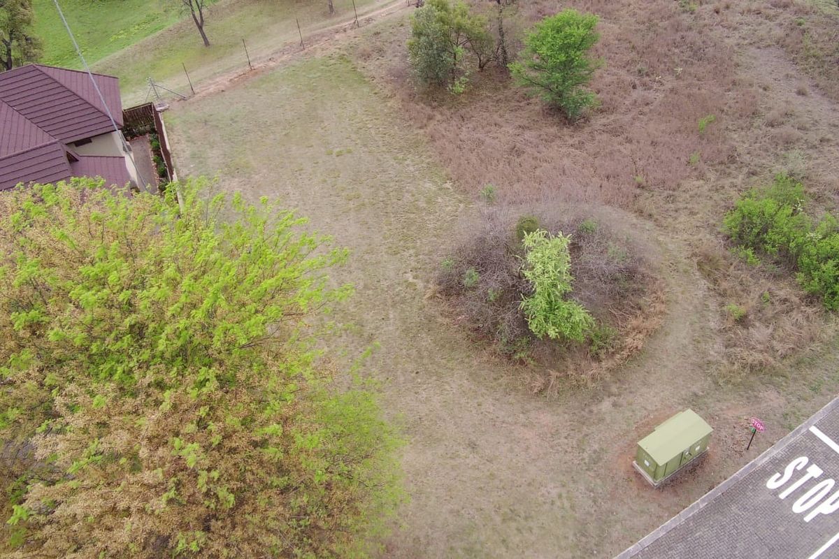 Aerial view of a vacant residential plot featuring natural vegetation, mature trees, perimeter fencing, and adjacent paved road access.