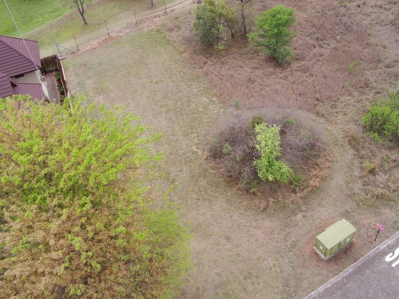 Aerial view of a vacant residential plot featuring natural vegetation, mature trees, perimeter fencing, and adjacent paved road access.