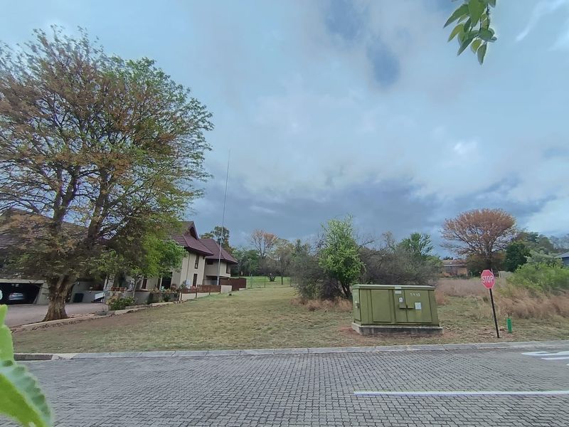 A view of a vacant residential plot from the street, featuring mature trees and an electrical transformer box on the property boundary.