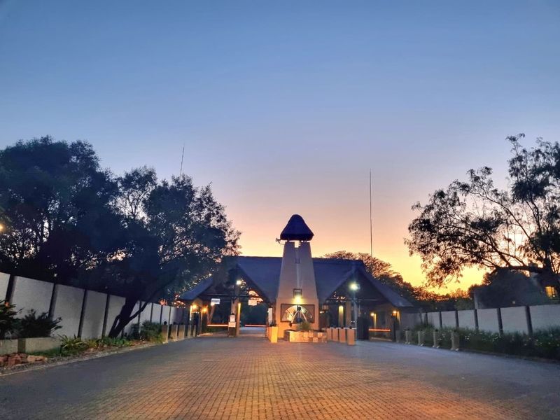 A well-maintained gated residential entrance featuring a paved driveway, security guardhouse, and perimeter wall at sunset.
