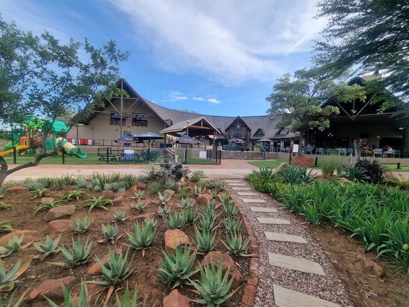 A well-maintained clubhouse facility featuring a thatched roof, expansive outdoor seating areas, and beautifully landscaped gardens with a stone-paved pathway.