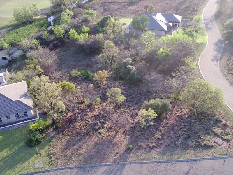 An aerial view of a vacant residential plot situated within a developed estate, featuring established trees and proximity to paved roads and neighboring homes.