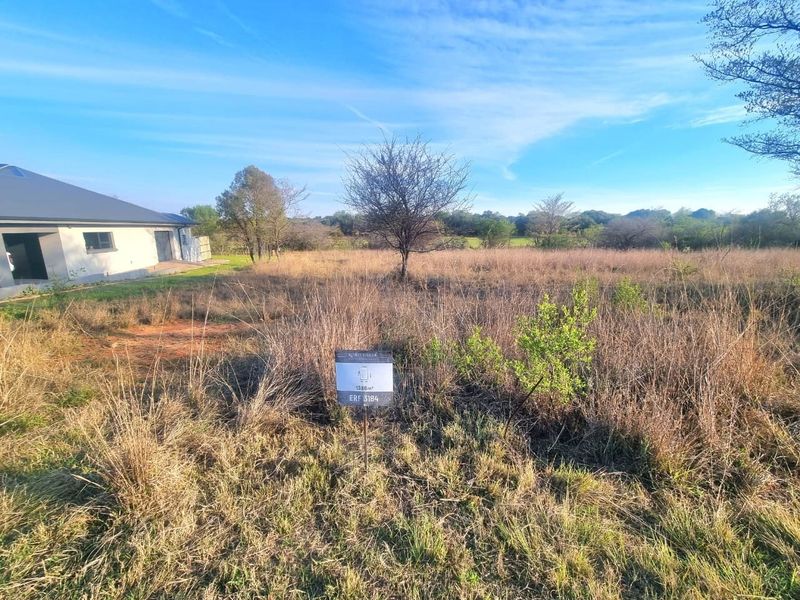 A view of a vacant residential plot featuring natural vegetation and property signage, situated adjacent to an existing residential structure.