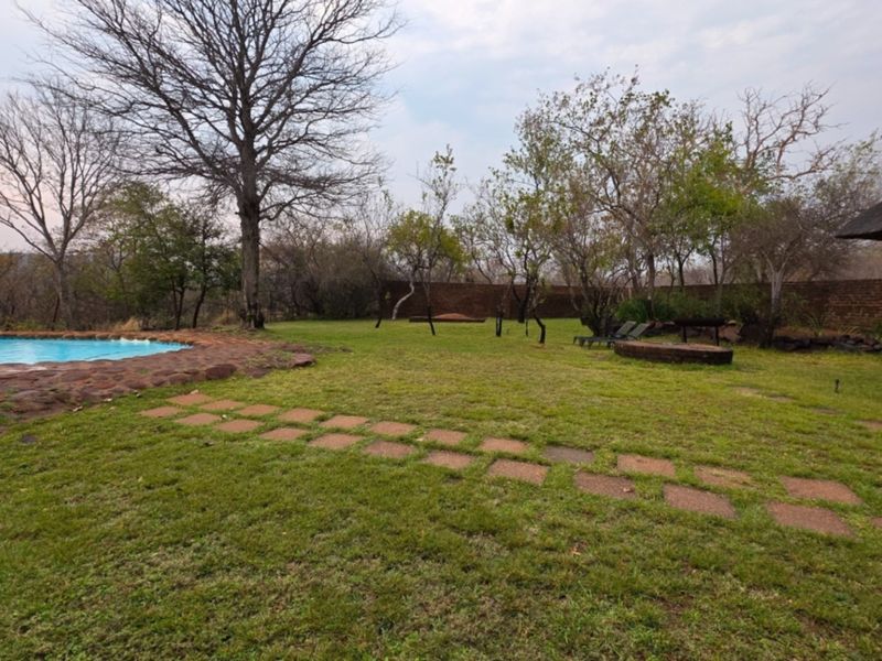 Spacious garden area featuring a swimming pool with stone coping, a thatched lapa, and a brick boundary wall, set within a natural landscape.