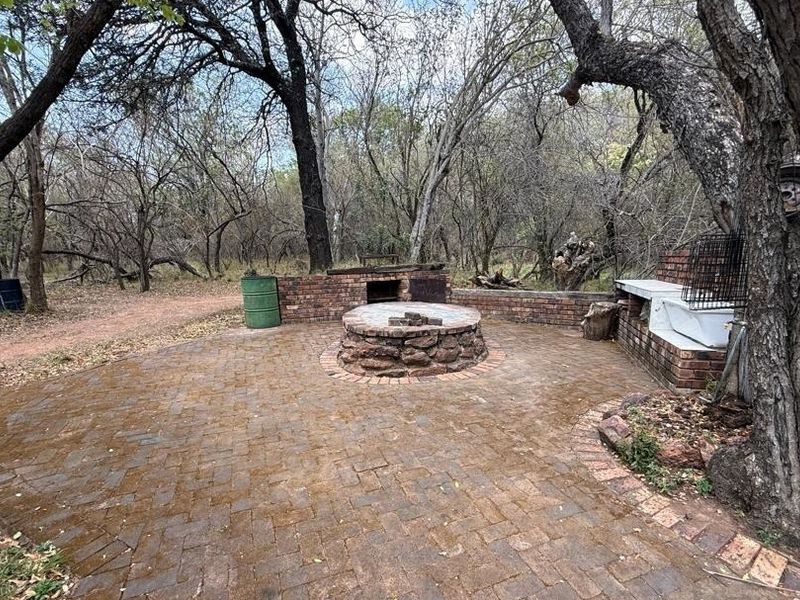 Outdoor entertainment area featuring a brick-paved patio, built-in braai, and a central stone fire pit set within a natural wooded environment.